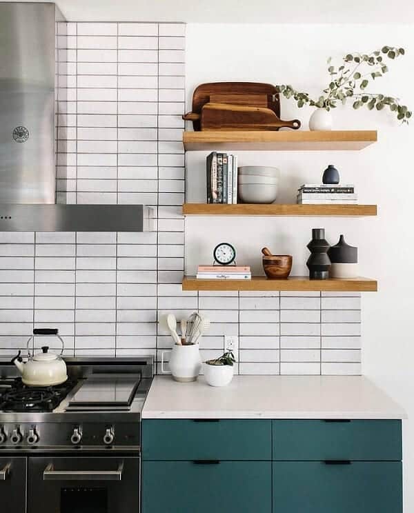 Cocina con placa para salpicaduras de azulejos blancos apilados, gabinetes verdes y estantes flotantes de madera