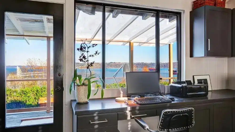 Escritorio de oficina frente a las ventanas y una puerta de vidrio, que deja pasar la luz del sol y muestra una vista natural.
